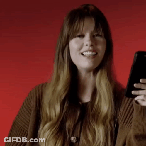 A Woman With Long Hair Is Smiling While Holding A Cell Phone . GIF
