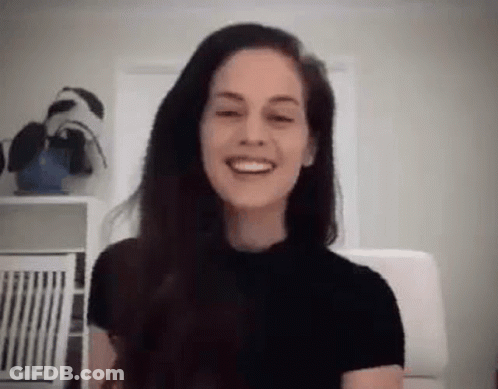 A Woman With Long Hair Is Smiling While Sitting In A Chair In A Room . GIF