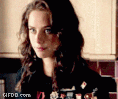 A Woman With Long Hair Is Standing In A Kitchen Wearing A Black Sweater With Stickers On It . GIF