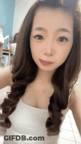 A Woman With Long Hair Is Taking A Selfie In A White Tank Top GIF