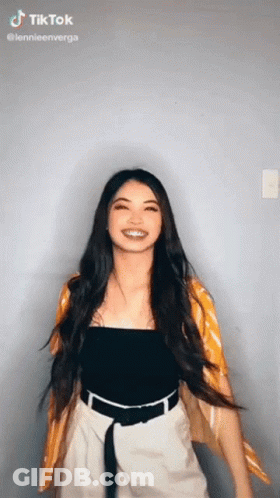 A Woman With Long Hair Is Wearing A Black Top And White Shorts And A Yellow Cardigan . GIF