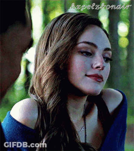 A Woman With Long Hair Is Wearing A Blue Sweater And A Necklace . GIF