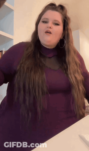 A Woman With Long Hair Is Wearing A Purple Turtleneck And Hoop Earrings GIF