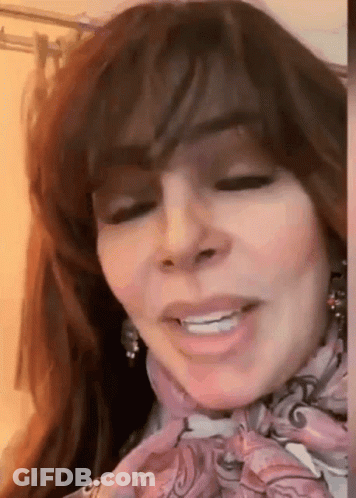 A Woman With Long Hair Is Wearing A Scarf Around Her Neck And Smiling . GIF