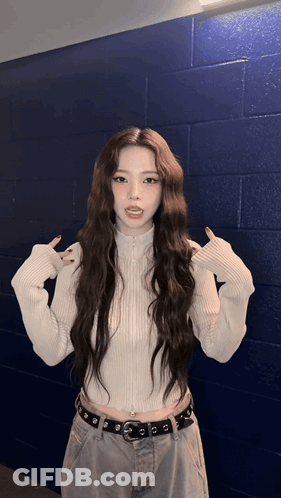 A Woman With Long Hair Wearing A White Sweater And Jeans Stands In Front Of A Blue Brick Wall GIF