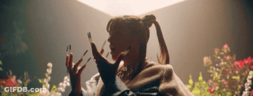 A Woman With Long Nails And A Ponytail Is Standing In Front Of Flowers . GIF