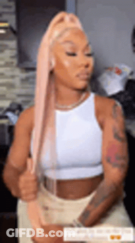 A Woman With Long Pink Hair Is Wearing A White Tank Top And Tan Shorts . GIF
