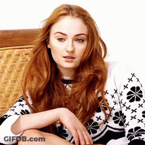 A Woman With Long Red Hair Is Sitting On A Wicker Chair Wearing A Black And White Sweater With Flowers On It . GIF