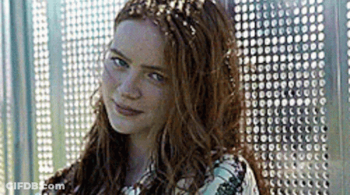 A Woman With Long Red Hair Is Standing In Front Of A Metal Wall And Smiling . GIF