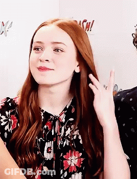 A Woman With Long Red Hair Is Wearing A Floral Shirt And Waving . GIF