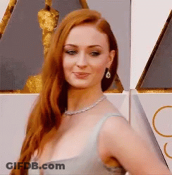 A Woman With Long Red Hair Wearing A Necklace And Earrings Stands In Front Of An Oscar Statue GIF