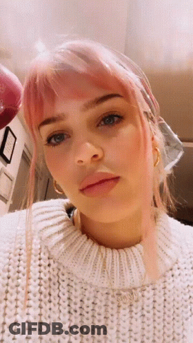 A Woman With Pink Hair And A White Sweater Looks At The Camera GIF