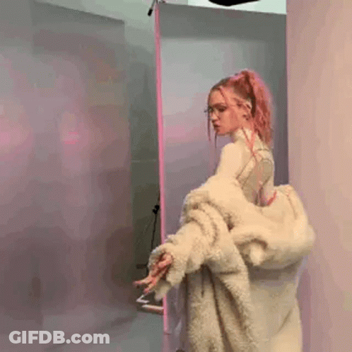 A Woman With Pink Hair And Glasses Is Wearing A Fur Coat And Standing In A Room . GIF