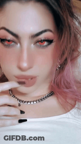 A Woman With Pink Hair And Red Eyes Is Wearing A White Shirt And A Chain Around Her Neck . GIF