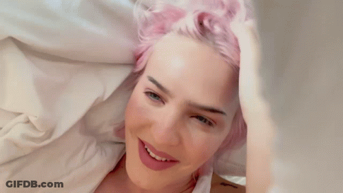 A Woman With Pink Hair Is Laying On A Bed And Smiling GIF