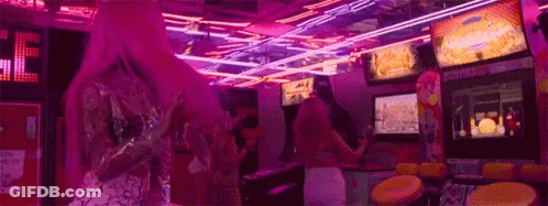 A Woman With Pink Hair Is Playing A Video Game In An Arcade With Neon Lights . GIF
