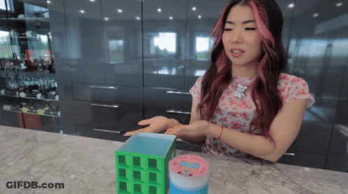 A Woman With Pink Hair Is Sitting At A Counter Holding A Container That Says ' Hello Kitty ' On It GIF
