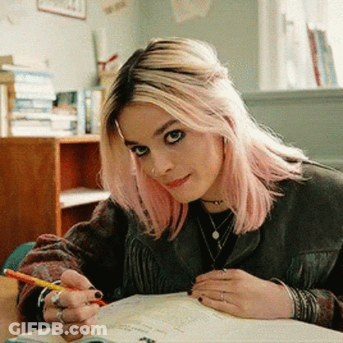 A Woman With Pink Hair Is Sitting At A Desk With A Pencil In Her Hand GIF
