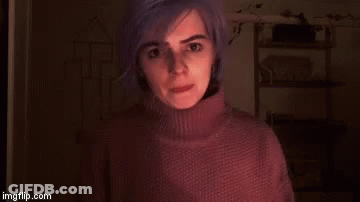 A Woman With Purple Hair Is Wearing A Turtleneck Sweater And Standing In A Dark Room . GIF