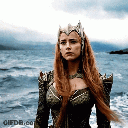 A Woman With Red Hair And A Crown On Her Head Stands In Front Of A Body Of Water GIF