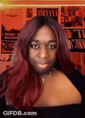 A Woman With Red Hair And A Necklace With A Cross In Front Of A Nirvana Poster GIF