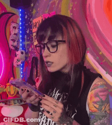 A Woman With Red Hair And Glasses Is Sitting At A Table Holding A Cell Phone . GIF