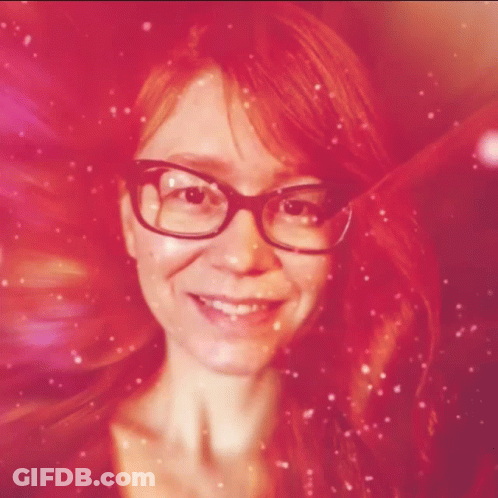 A Woman With Red Hair And Glasses Smiles In A Pink Background GIF