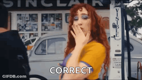 A Woman With Red Hair Is Blowing A Kiss While Standing In Front Of A Gas Pump And A Car . GIF