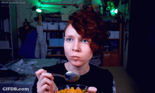 A Woman With Red Hair Is Eating A Bowl Of Food With A Spoon In Front Of A Sign That Says N7 On It GIF