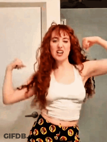 A Woman With Red Hair Is Flexing Her Muscles In Front Of A Door . GIF