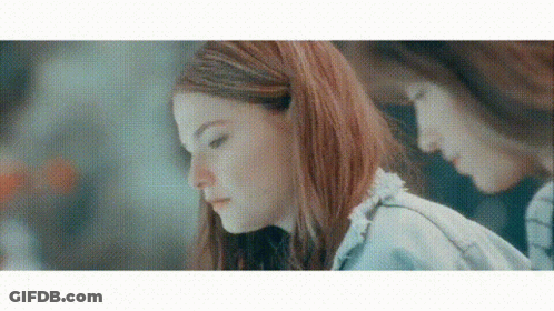A Woman With Red Hair Is Looking Down At Something While Another Woman Looks At Her Reflection In The Mirror . GIF