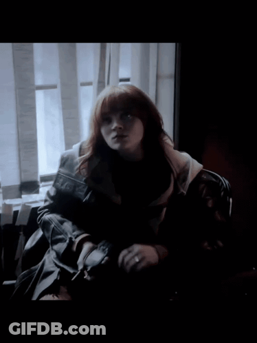 A Woman With Red Hair Is Sitting In Front Of A Window In A Dark Room GIF