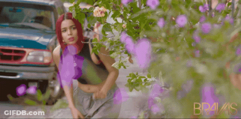 A Woman With Red Hair Is Standing In Front Of A Bush With Purple Flowers And The Word Bravis On The Bottom Right GIF