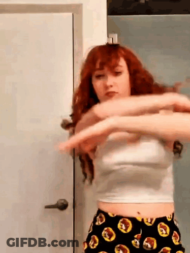 A Woman With Red Hair Is Standing In Front Of A Door Wearing A White Crop Top And Black Shorts . GIF