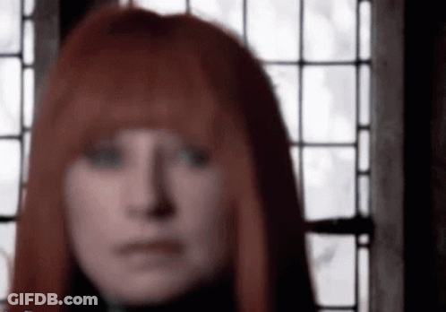 A Woman With Red Hair Is Standing In Front Of A Window And Looking At The Camera . GIF