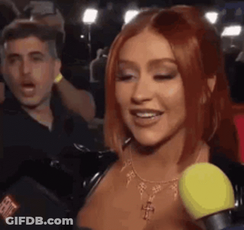 A Woman With Red Hair Is Talking Into A Microphone While A Man Looks On . GIF