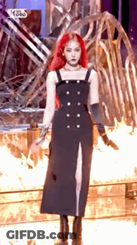 A Woman With Red Hair Is Wearing A Black Dress And Standing In Front Of A Fire . GIF