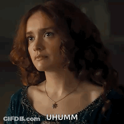 A Woman With Red Hair Is Wearing A Necklace And A Blue Dress And Looking At The Camera . GIF