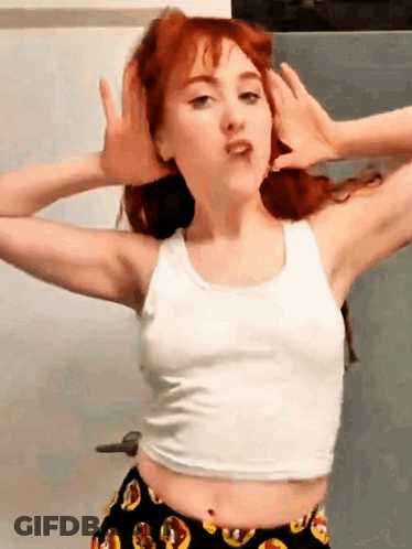 A Woman With Red Hair Is Wearing A White Tank Top And Black Shorts GIF
