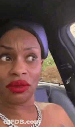 A Woman With Red Lipstick And A Necklace Is Sitting In A Car Making A Funny Face GIF