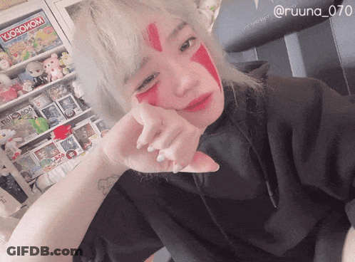 A Woman With Red Paint On Her Face Is Standing In Front Of A Shelf With A Monopoly Game GIF