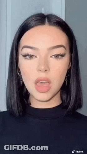 A Woman With Short Black Hair And Earrings Is Wearing A Black Sweater And Making A Funny Face . GIF
