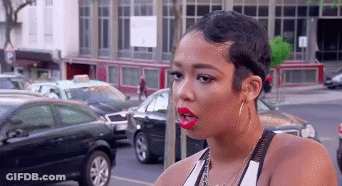 A Woman With Short Hair And Red Lipstick Is Standing On A Street . GIF