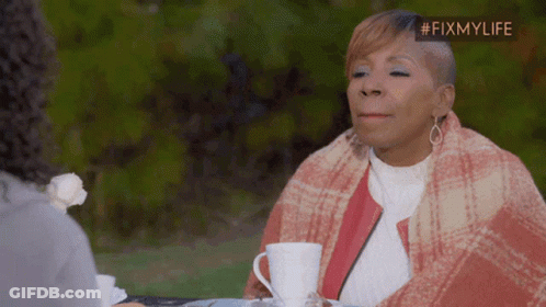 A Woman Wrapped In A Plaid Blanket Sits At A Table With A Cup Of Coffee In Front Of Her GIF