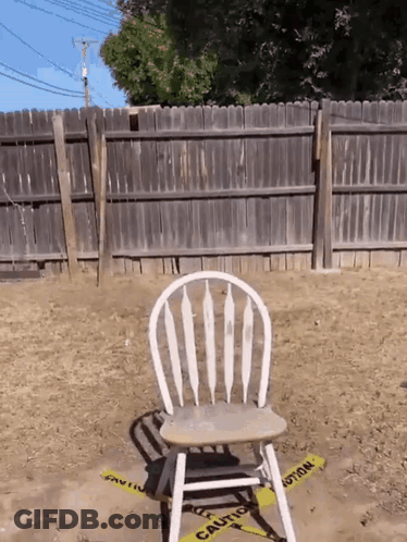 A Wooden Chair Sits In The Dirt In Front Of A Fence With A Yellow Caution Tape Around It GIF