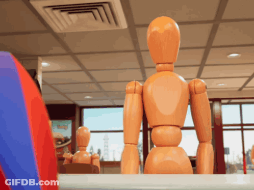 A Wooden Mannequin Sits At A Table In A Room GIF