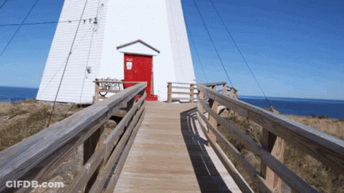 A Wooden Walkway Leads To A White Building With A Red Door And A Sign That Says No Dogs GIF