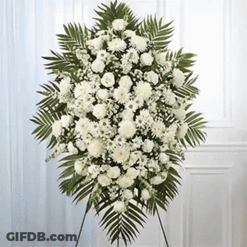 A Wreath Of White Flowers And Green Leaves Is Sitting On A Stand In Front Of A Door . GIF