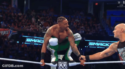 A Wrestler In A Green Outfit Holds Hands With Another Wrestler In Front Of A Smack Club Sign GIF