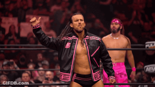 A Wrestler In A Pink And Black Jacket Stands In A Ring With Two Other Men GIF
